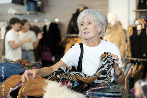 Happy mature woman looking at second hand clothes in large clothing shop