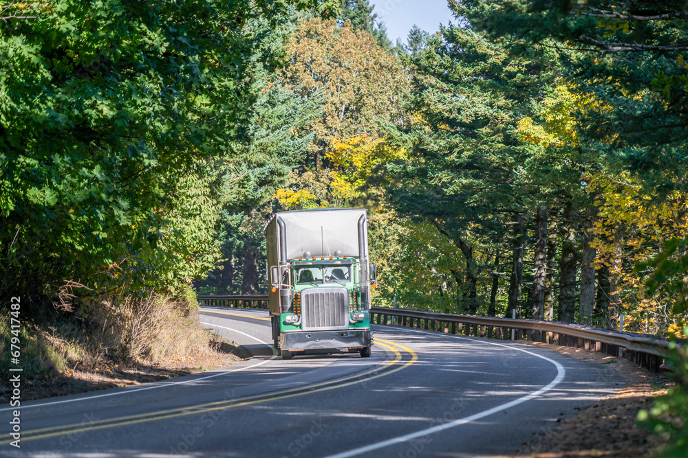 Classic green big rig semi truck tractor with high exhaust pipes transporting commercial cargo in bulk semi trailer driving on the winding narrow road in autumn forest