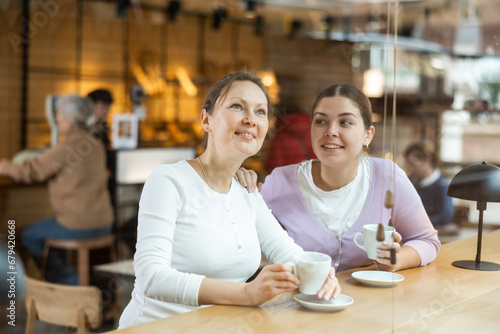 Portrait of two joyful European women joking with each other while sitting with cups of coffee in cafe