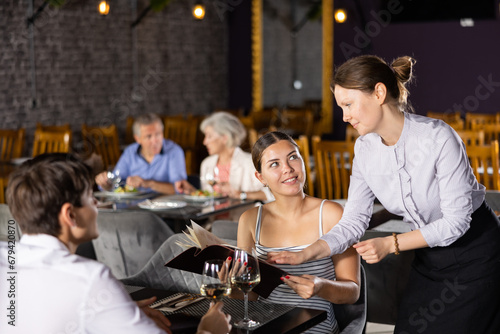 Young male and female colleagues having nice time in sparsely populated cozy restaurant. Waitress answers questions of visitors, helps to choose diet dish for late dinner in menu photo