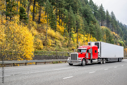 Red classic long hauler American big rig semi truck transporting frozen cargo in loaded reefer semi trailer driving on the autumn highway road in Columbia River Gorge