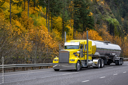 Yellow bright classic big rig semi truck tractor transporting liquid cargo in tank semi trailer running on the picturesque autumn road with rock mountain on the side photo