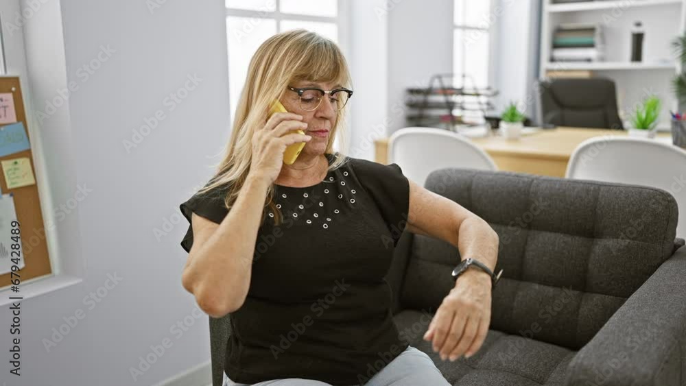 Elegant blonde middle-aged woman boss, immersed in a serious business talk over her smartphone, working indoors at office workplace. Elegant blonde middle-aged woman boss, immersed in a serious business talk over her smartphone, working indoors at office workplace.