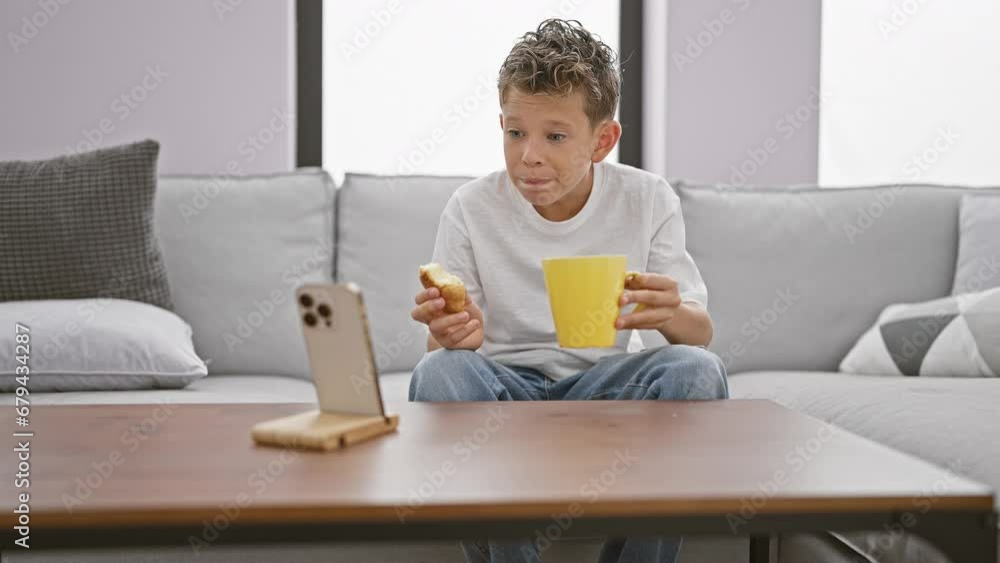 Adorable little blond boy, engrossed in watching video on smartphone, enjoys breakfast cake snack sitting on sofa at home in the living room Adorable little blond boy, engrossed in watching video on smartphone, enjoys breakfast cake snack sitting on sofa at home in the living room