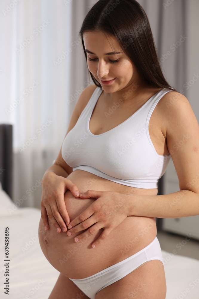 Beautiful pregnant woman in stylish comfortable underwear making heart with hands on her belly indoors