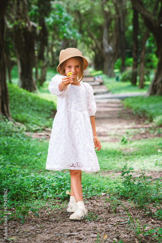 A little girl in a dark forest on a path gives a flower. A girl in a white dress and hat