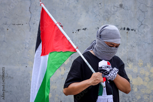Man wearing a keffiyeh holds the Palestinian flag with hand on chest.