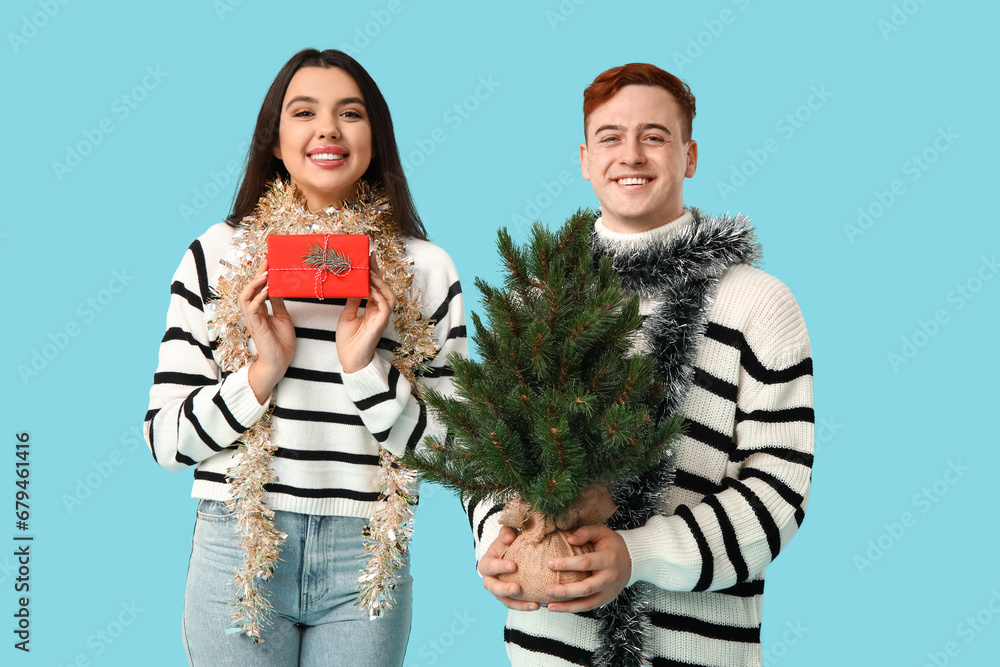 Young couple with Christmas tree, tinsel and gift box on blue background Young couple with Christmas tree, tinsel and gift box on blue background
