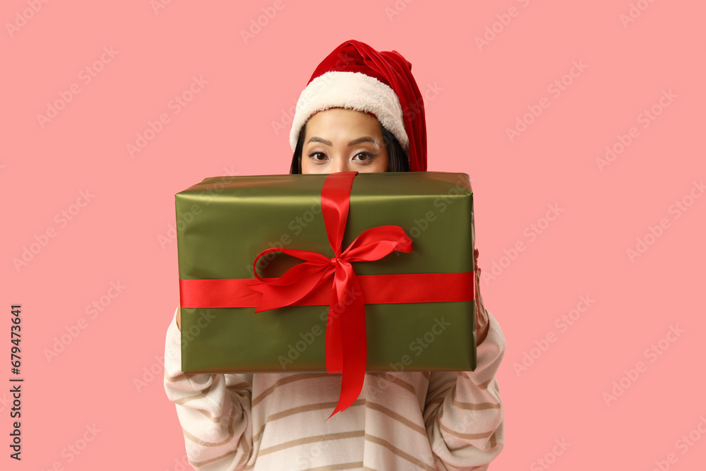 Young Asian woman in Santa hat with Christmas gift box on pink background Young Asian woman in Santa hat with Christmas gift box on pink background