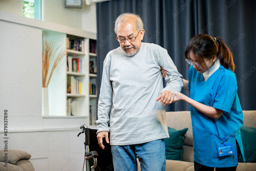 Fototapeta premium Asian doctor support old man to getting up to exercise, help handicapped elderly stand up, woman nurse assisting helping senior man patient get up from wheelchair for practice walking at home