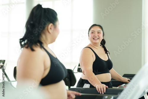 Two Asian women with chubby bodies, Let's meet at fitness center where we often exercise, greet each other until we get along, standing, talking in front treadmill, happy expressions.