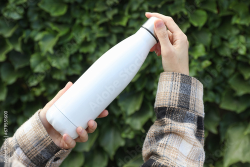 Hand holds white thermos bottle on green leaves wall background