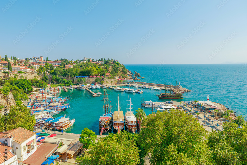 Naklejka premium Panoramic view of Antalya, Turkey. Deep blue-green waters of the Mediterranean Sea meet a bustling harbor filled with boats of various sizes. A white lighthouse stands sentinel on a rocky outcropping