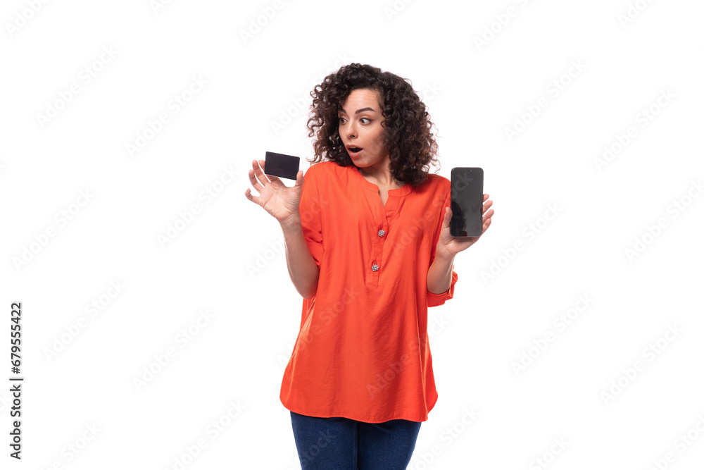 young caucasian brunette woman with curly hair dressed in an orange blouse demonstrates in surprise a plastic card and a smartphone young caucasian brunette woman with curly hair dressed in an orange blouse demonstrates in surprise a plastic card and a smartphone