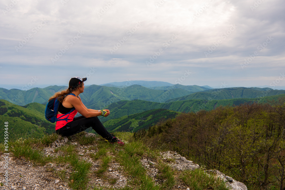 Young woman with backpack sitting on the mountain peak and beautiful mountains . Landscape with sporty girl, green grass, forest, hills, sky, sunbeams in fall. Travel. Nature