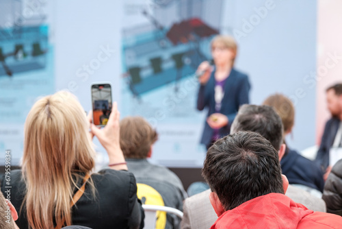 Businesswoman photographing speaker through smart phone while attending global presentation in conference hall