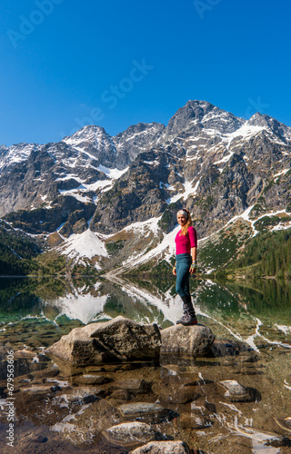 Young woman in red sportswear standing in the nature, mountain lake and range on the background, active life concept, summer time in Eye of the Sea lake in Tatra Mountains in Poland