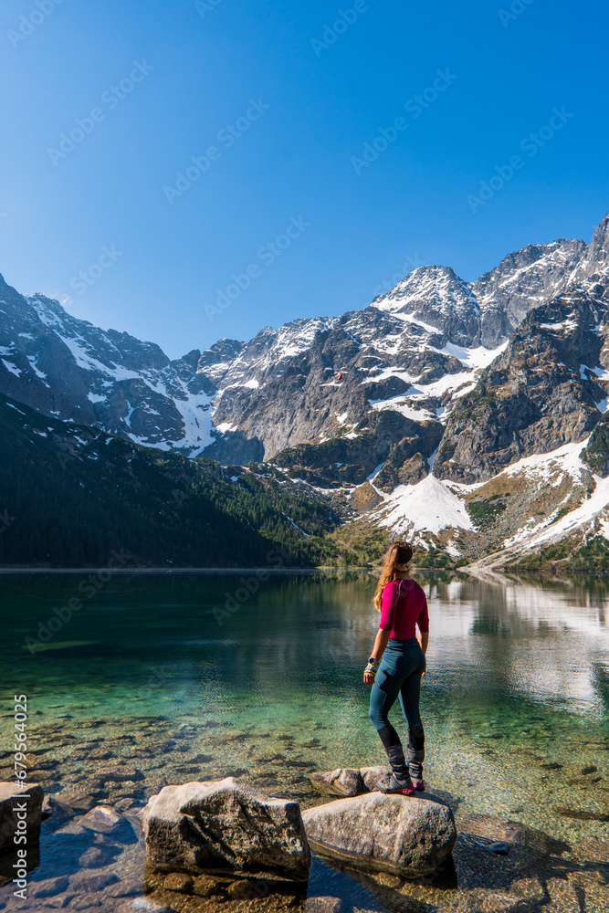 A tourist girl with a large backpack stands near an alpine lake in the stones in the lake Morskie oko or sea eye lake mountain against the backdrop of sea Eye and snow in autumn. - obrazy, fototapety, plakaty 