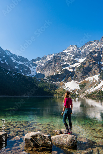 A tourist girl with a large backpack stands near an alpine lake in the stones in the lake Morskie oko or sea eye lake mountain against the backdrop of sea Eye and snow in autumn. photo