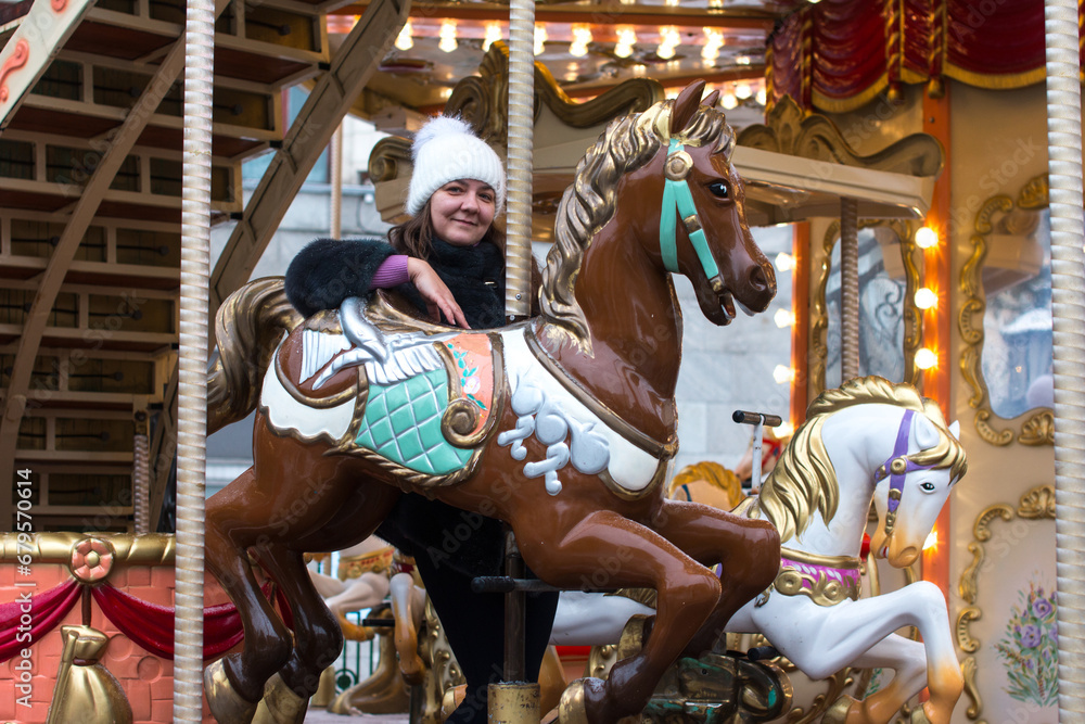 A woman in a winter fur coat rides a carousel in the winter park on the square. New Year's walk around the square