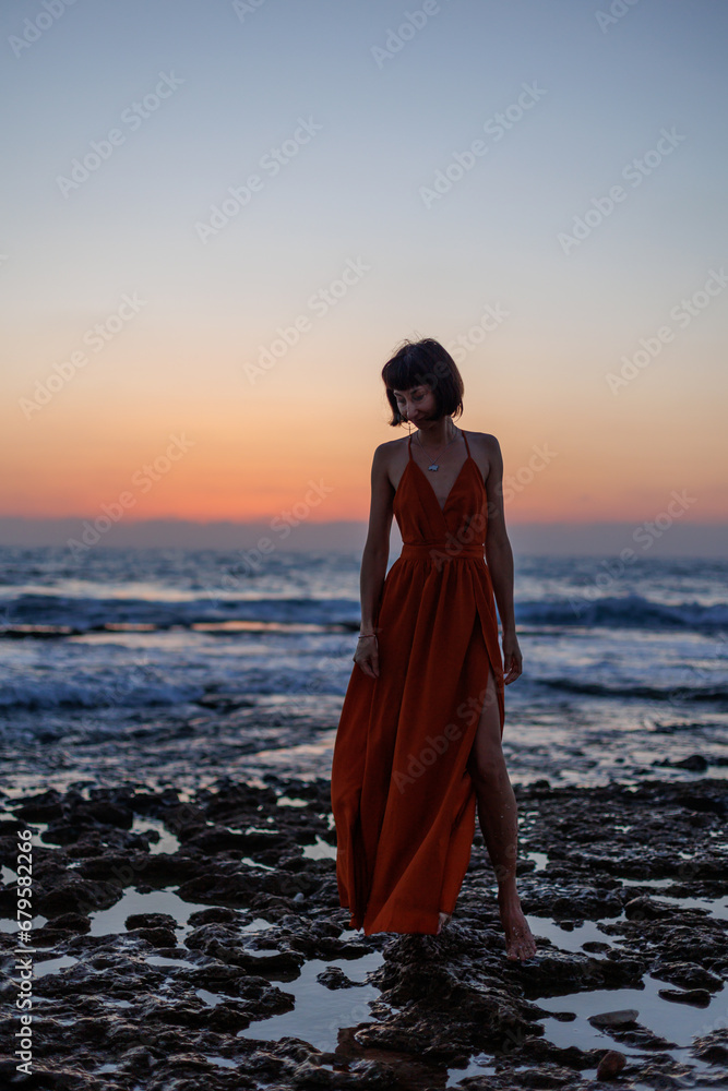 young woman in a summer dress walks along the seashore during sunset and looks at the sea. young woman in a summer dress walks along the seashore during sunset and looks at the sea.