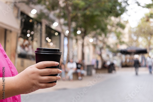 A close-up of a woman holding a black plastic coffee cup in hand. Coffee break in the city center with a bustling cityscape in the background on a sunny summer day.