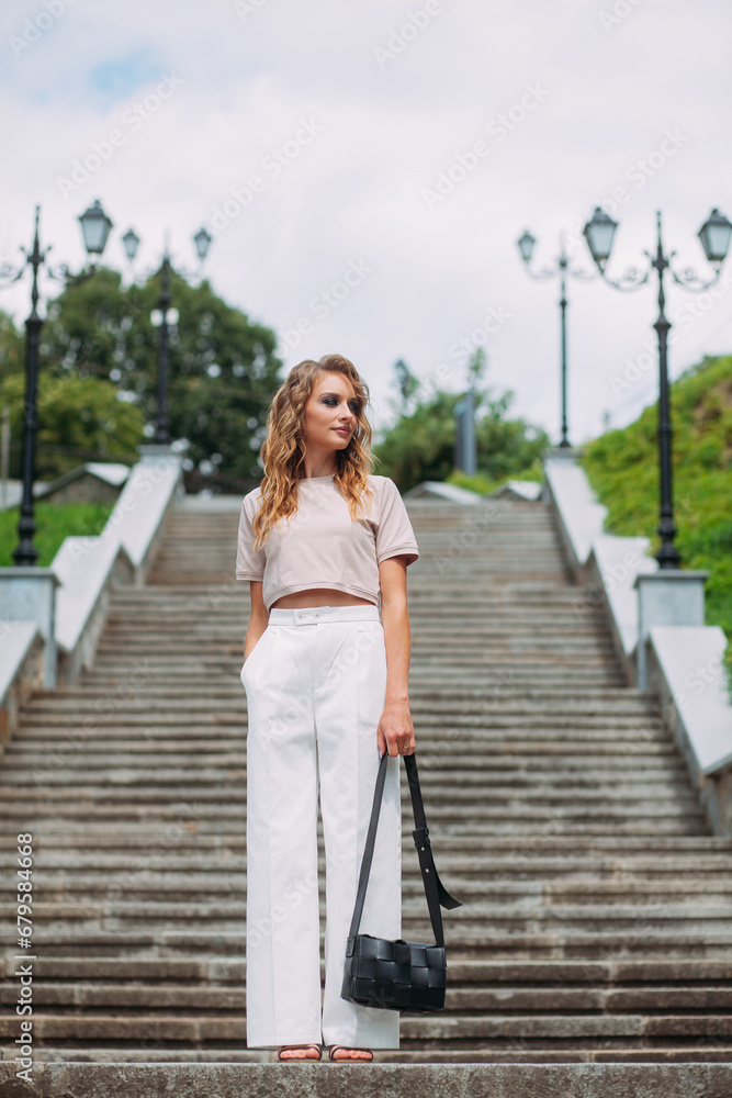 A stylish girl with fashionable makeup and hairstyle, in white trousers and a beige top and a handbag in her hands, stands against the backdrop of a large stone staircase in the park.