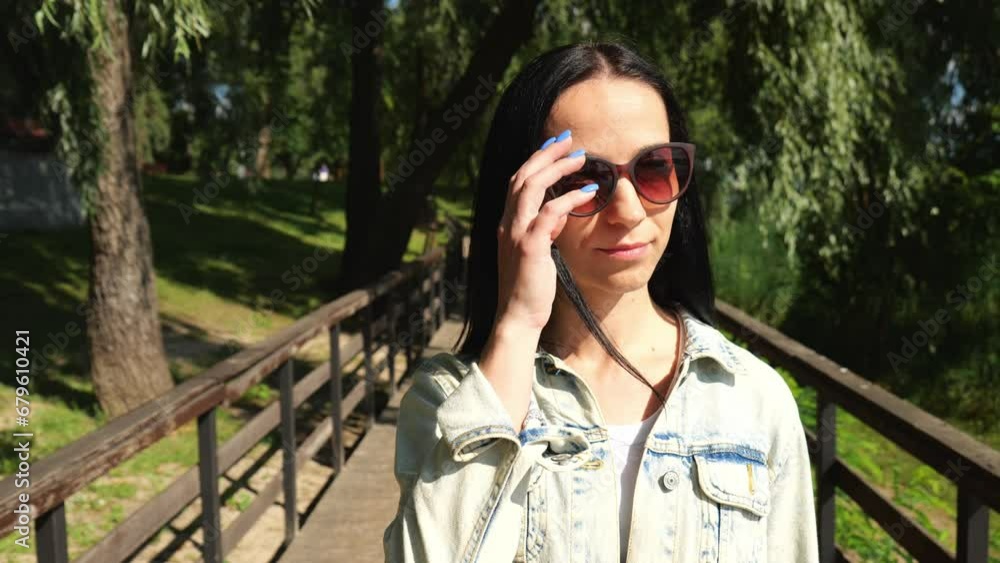 Portrait of a young brunette woman in sunglasses walks along a wooden bridge in a summer park. Slow motion portrait of a beautiful woman. Woman in a denim jacket Portrait of a young brunette woman in sunglasses walks along a wooden bridge in a summer park. Slow motion portrait of a beautiful woman. Woman in a denim jacket