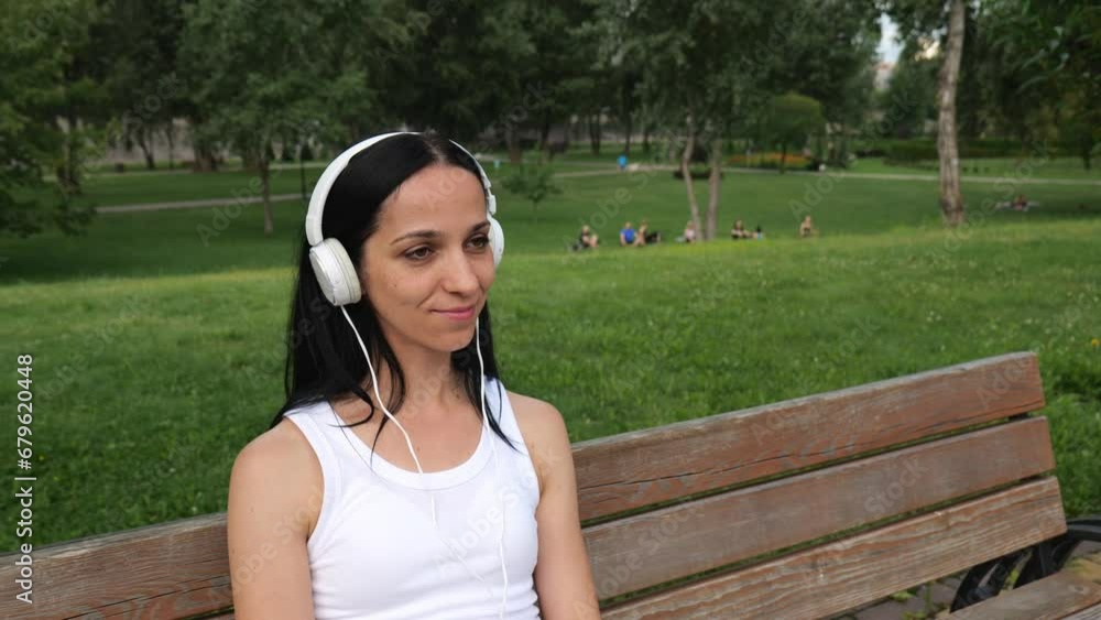 Portrait of a young brunette woman with white headphones sitting on a bench in a summer park. A thirty-year-old woman listens to music on headphones. slow motion portrait woman music Portrait of a young brunette woman with white headphones sitting on a bench in a summer park. A thirty-year-old woman listens to music on headphones. slow motion portrait woman music