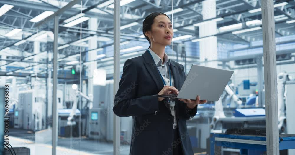 Asian Factory Manager Working on Laptop at a Futuristic Industrial Manufacture with Autonomous Robotic Assembly Line. Specialist Monitoring Production, Planning Work for Employees Asian Factory Manager Working on Laptop at a Futuristic Industrial Manufacture with Autonomous Robotic Assembly Line. Specialist Monitoring Production, Planning Work for Employees