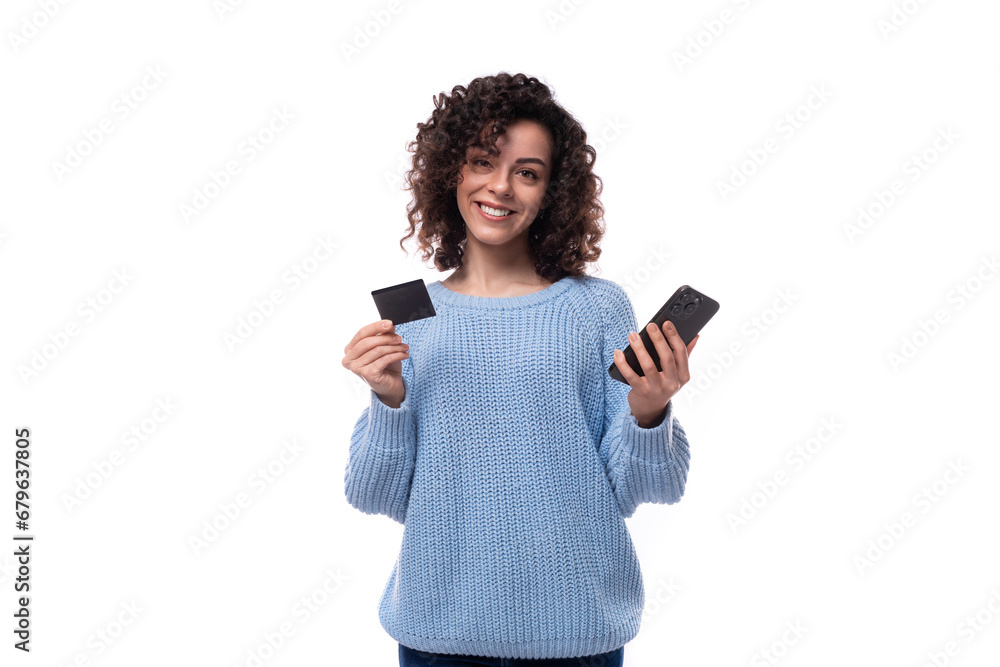 authentic smiling young brunette woman with curls is dressed in a blue knitted sweater holding a plastic card and a smartphone. e-business concept authentic smiling young brunette woman with curls is dressed in a blue knitted sweater holding a plastic card and a smartphone. e-business concept