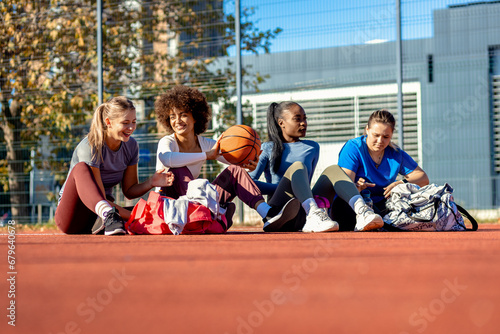 Diverse group of young woman sitting on court resting afrer playing basketball outdoors. photo