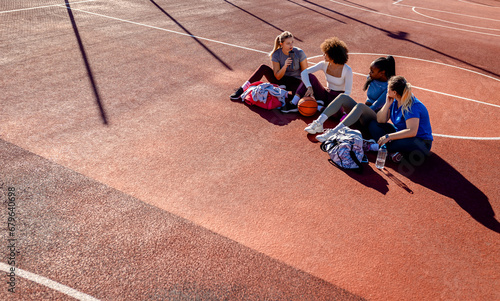 Diverse group of young woman sitting on court resting afrer playing basketball outdoors. photo