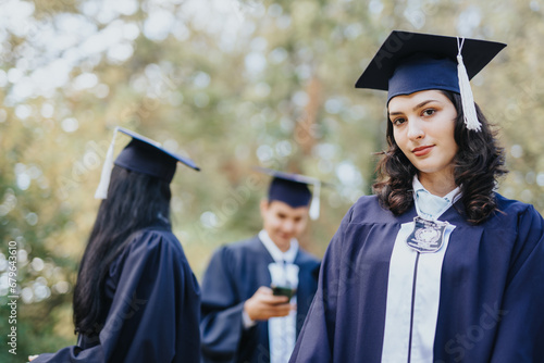 Portrait of graduated, university, female student wearing gown and cap looking at the camera. Students discussing in the back. Copy space.