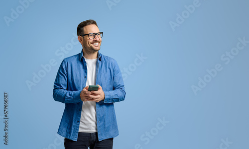 Smiling businessman holding mobile phone and looking away thoughtfully against blue background