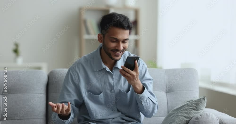 Cheerful young Indian guy talking on video conference call on smartphone, holding cellphone, speaking, smiling, enjoying online conversation, looking at screen, sitting on soft home couch Cheerful young Indian guy talking on video conference call on smartphone, holding cellphone, speaking, smiling, enjoying online conversation, looking at screen, sitting on soft home couch