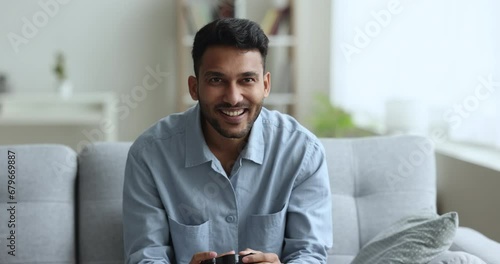 Cheerful young Indian gamer guy playing virtual game at home, holding joystick, swinging on home couch, pushing buttons on gamepad device, smiling, laughing, getting excited photo