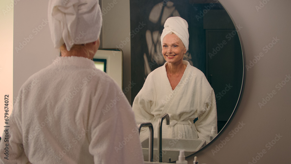 Obraz premium Female beauty spa procedure anti-aging health skincare middle-aged mature woman in bathrobe after shower rejuvenation hydrated skin beautiful Caucasian old lady smiling looking at mirror home bathroom