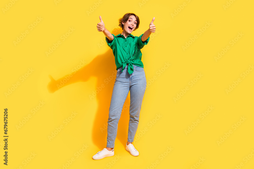 Full length photo of cool confident girl dressed green shirt smiling showing you thumbs up isolated yellow color background Full length photo of cool confident girl dressed green shirt smiling showing you thumbs up isolated yellow color background