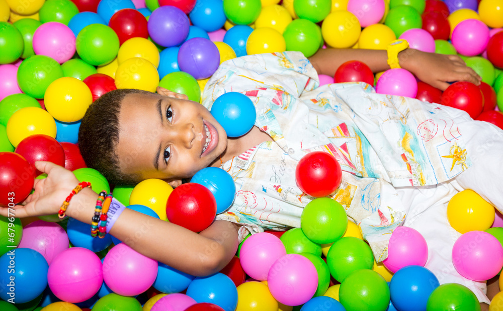Happy african black boy playing in ball pit on birthday party in kids amusement park and indoor play center. Child playing with colorful balls in playground ball pool.
