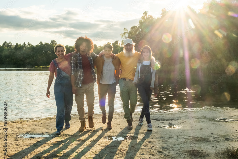 This high-quality photo captures a diverse group of millennial friends as they walk towards the camera along the forest lake shore. The warm glow of the setting sun envelops them, creating long