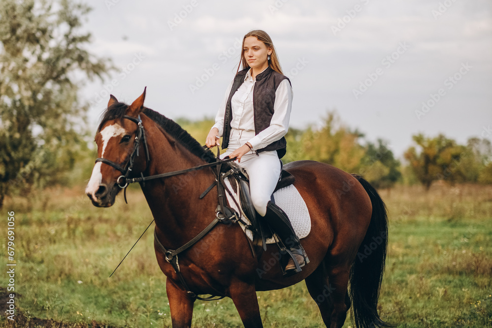 Fototapeta premium Young beautiful blonde woman jockey rides a brown horse in a meadow at sunset in summer. Preparing for an equestrian competition.