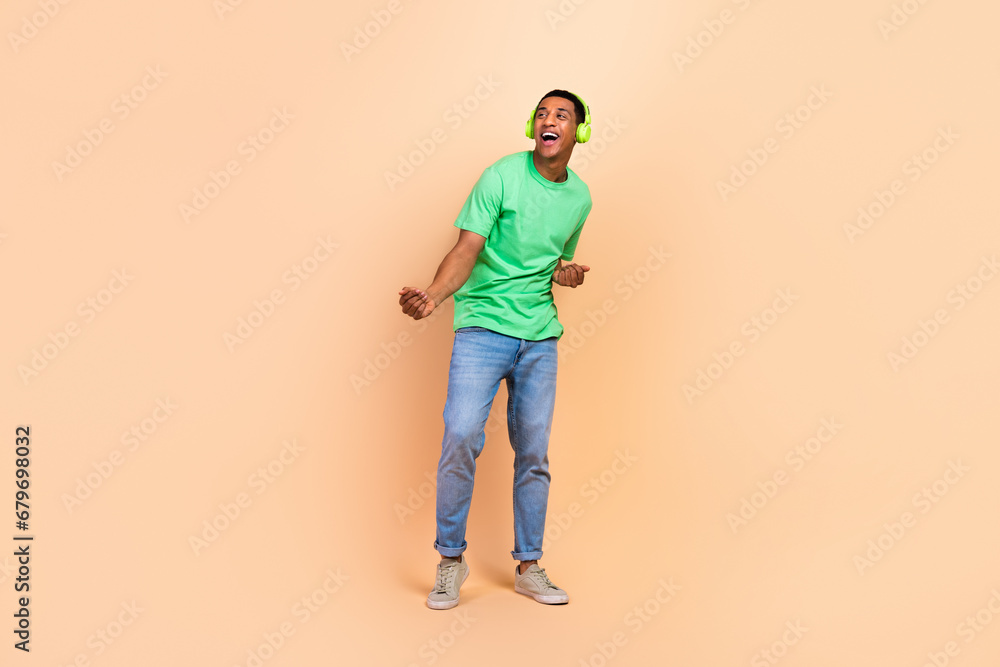 Full length photo of positive man wear stylish t-shirt jeans in headphones dancing look empty space isolated on beige color background