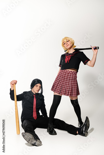 man in black wig sitting  with baseball bat near woman in school uniform on white, cosplay trend