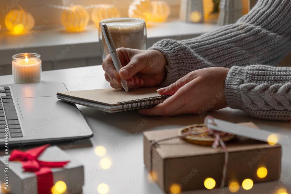 Fototapeta premium Young woman writing christmas cards, makes order on laptop. Female picking gifts online purchases at cozy home among gift boxes and packages. Winter sales, Black Friday. Christmas discount promotions