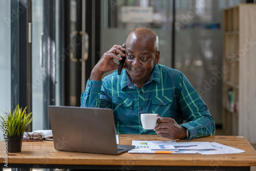 Senior black man talking on phone with attention tracking data on laptop screen. Sip coffee while talking on the phone.