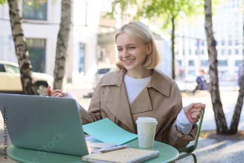 Portrait of young blond woman, modern female model sitting in outdoor cafe, drinking coffee, connects to online video chat via laptop, talking to coworkers remotely, gesturing and explaining smth photo