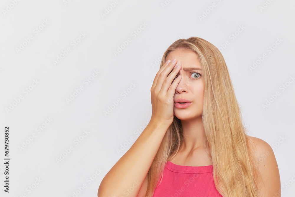 Young woman with long fair hair poses on white backdrop with one hand covering half of her beautiful face, hard day concept, copy space