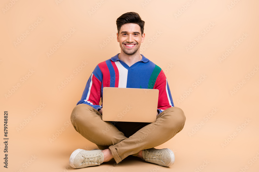 Full body length photo of positive it startuper young man working sit floor using his modern computer isolated on beige color background Full body length photo of positive it startuper young man working sit floor using his modern computer isolated on beige color background