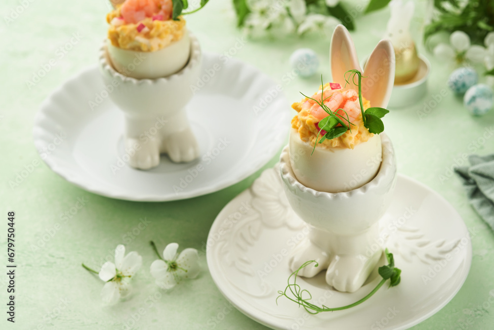 Stuffed or deviled eggs with yolk, shrimp, pea microgreens with paprika in rabbit-shaped stand for easter table decorate fresh cherry or apple blossoms on light background. Traditional dish for Easter Stuffed or deviled eggs with yolk, shrimp, pea microgreens with paprika in rabbit-shaped stand for easter table decorate fresh cherry or apple blossoms on light background. Traditional dish for Easter